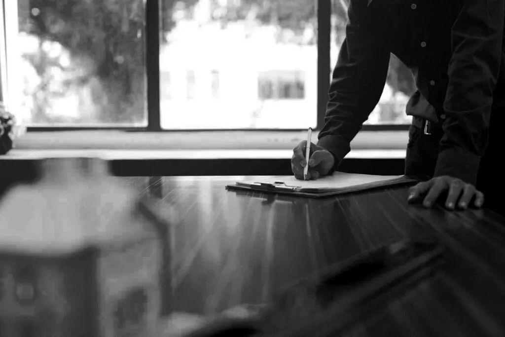 A person signing paperwork at a timber table beside a bright window