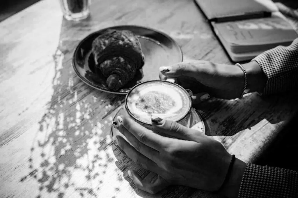 Hands cradling a cappuccino on a timber table beside a croissant