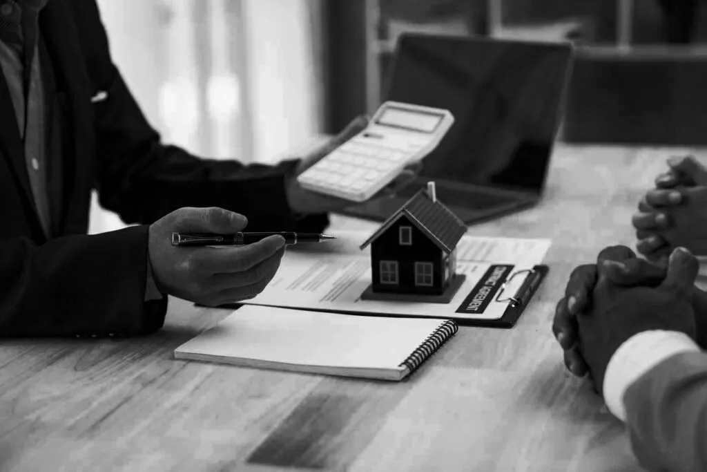Two people at a desk with a model house, calculator and notepad