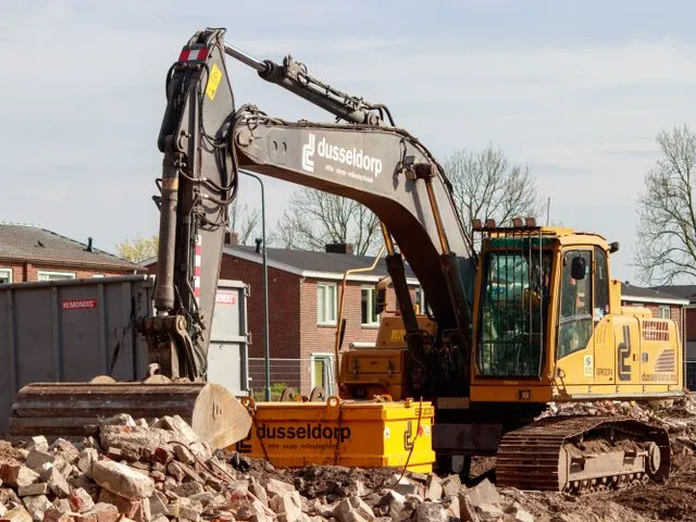 Demolition excavator clearing brick rubble in front of residential houses