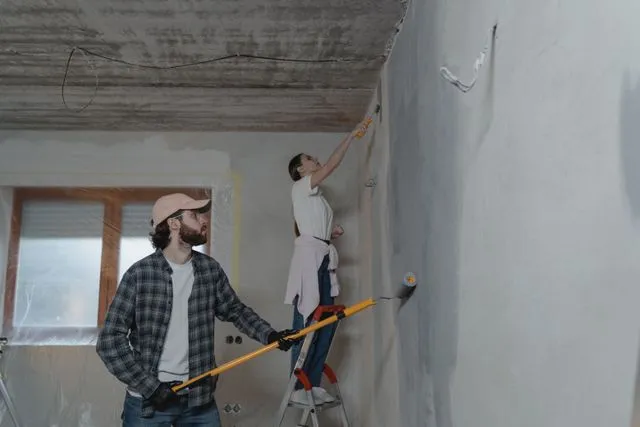 Two people renovating a room, one painting a wall with a roller and the other on a ladder
