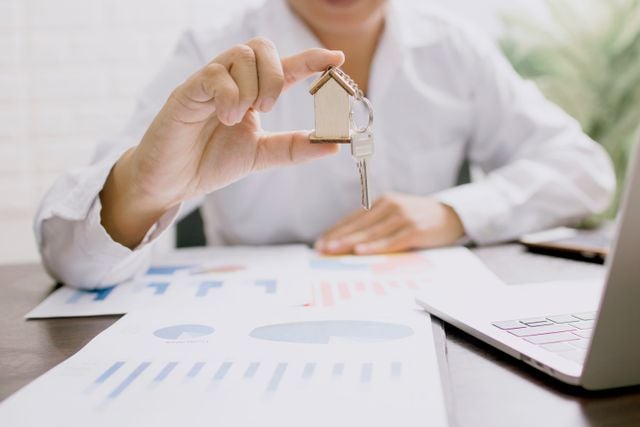 Hand holding a small house-shaped keyring above printed property market charts